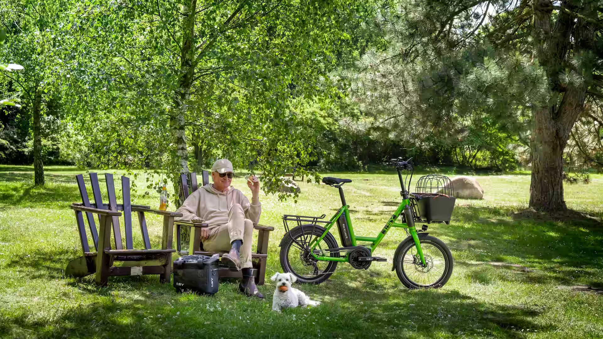 Froschgrünes i:SY S8k Kompaktrad mit MIK-Frontträger und Basil Buddy Hundekorb neben Herrchen im Holzstuhl und kleinem Hund im Park steigert i:SY Abenteuerlust
