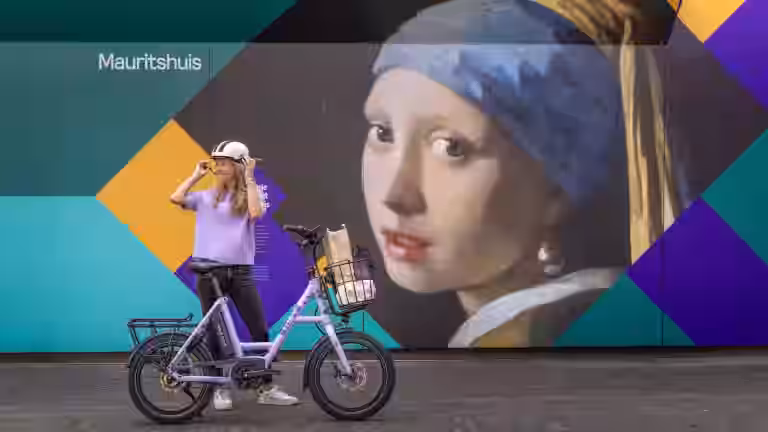 Eine stylische Fahrerin passt ihren ABUS HUD-Y Helm auf dem i:SY E5 in Pastel Purple vor dem Mauritshuis Wandgemälde an