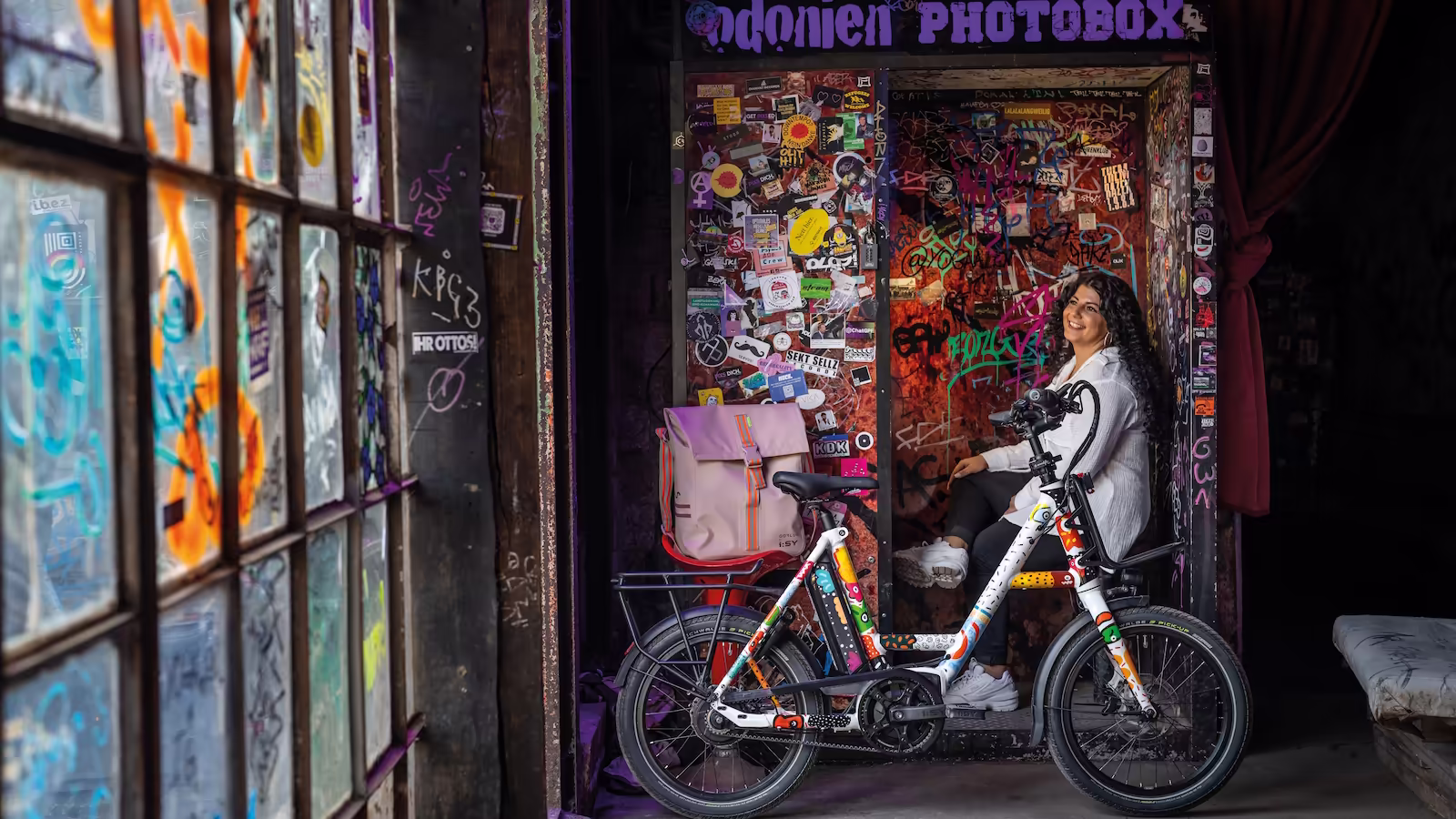 Yasemin sitzend in der Odonien-Photobox mit dem i:SY N3.8 ZR by Ona Sadkowsky E-Bike und der rosa Gofluo-Tasche davor im Farbenrausch